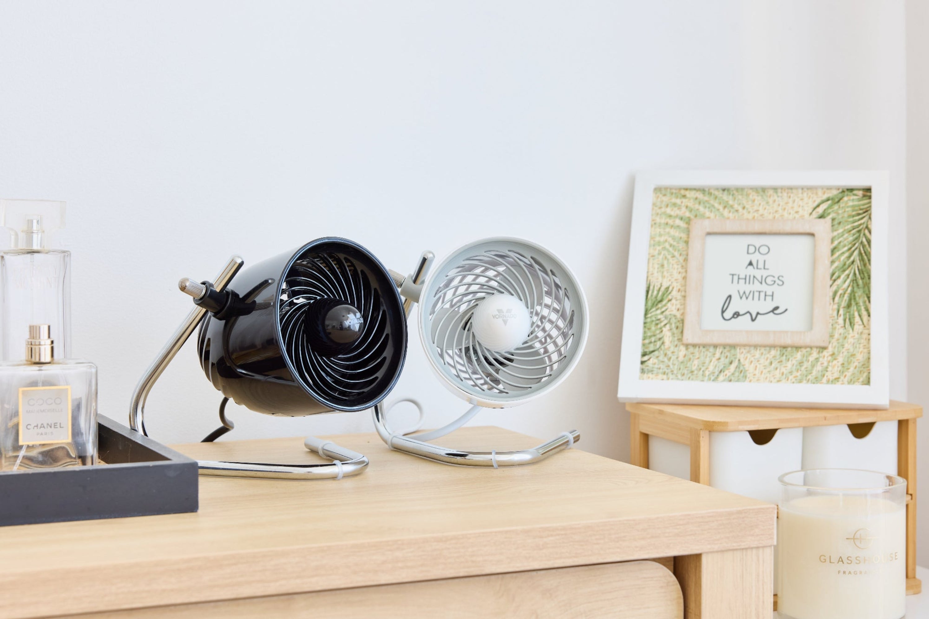 Two small Pivot desk fans side by side on wooden drawers with a framed picture in the background.