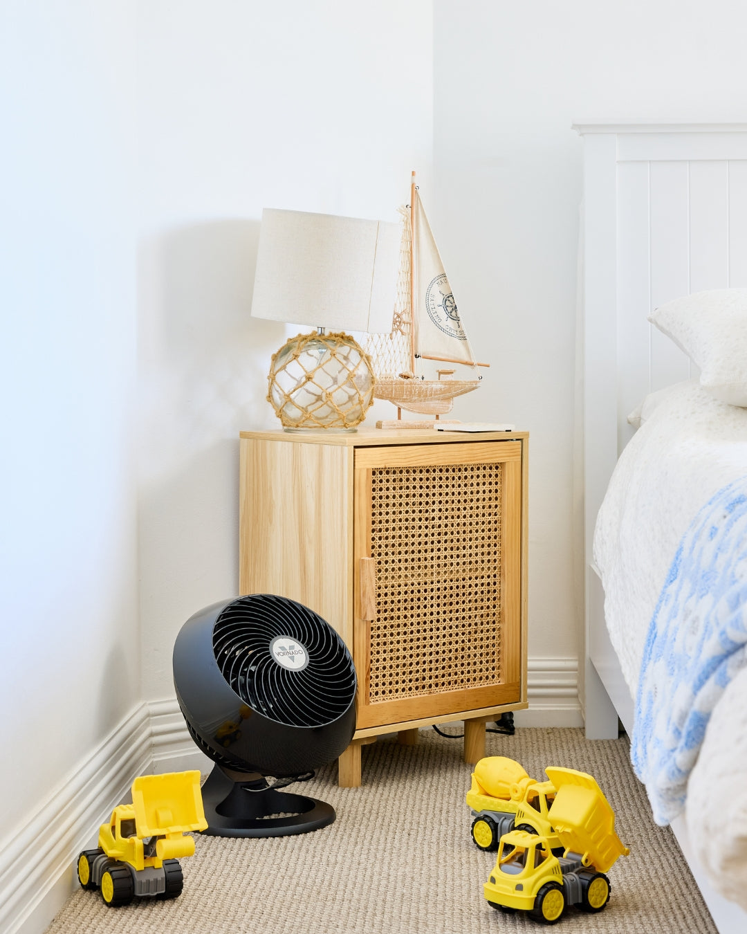 Black Vornado 460 Small Air Circulator on a carpeted floor of a bedroom, yellow toy trucks and a wooden cabinet with a lamp and boat model surround the Air Circulator.