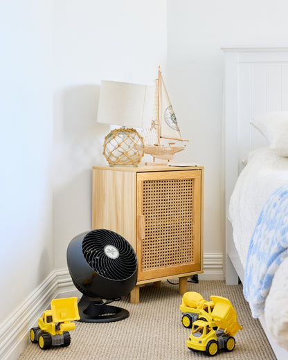 Black Vornado 460 Small Air Circulator on a carpeted floor of a bedroom, yellow toy trucks and a wooden cabinet with a lamp and boat model surround the Air Circulator.