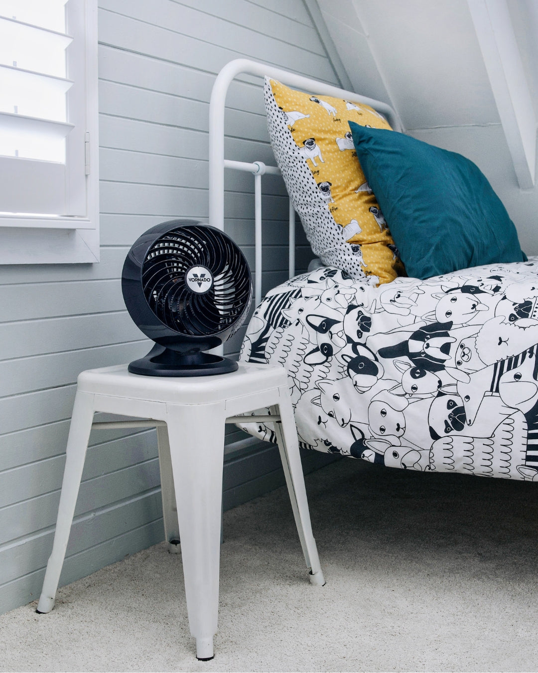 Black Vornado 460 Small Air Circulator on a white stool next to a bed with patterned bedding and pillows.