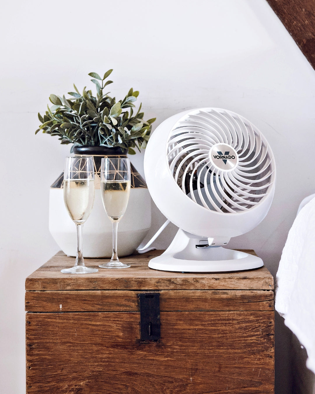 White Vornado 460 Small Air Circulator  on a wooden chest with two glasses of champagne and a plant in the background on beside a bed.
