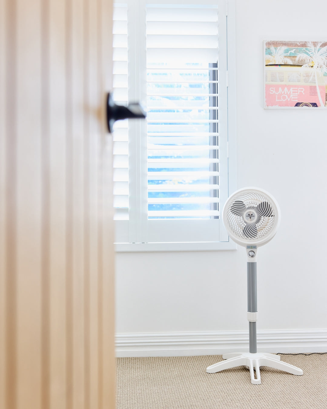 White Vornado 683 Pedestal Air Circulator on a carpeted floor with a window and curtain in the background