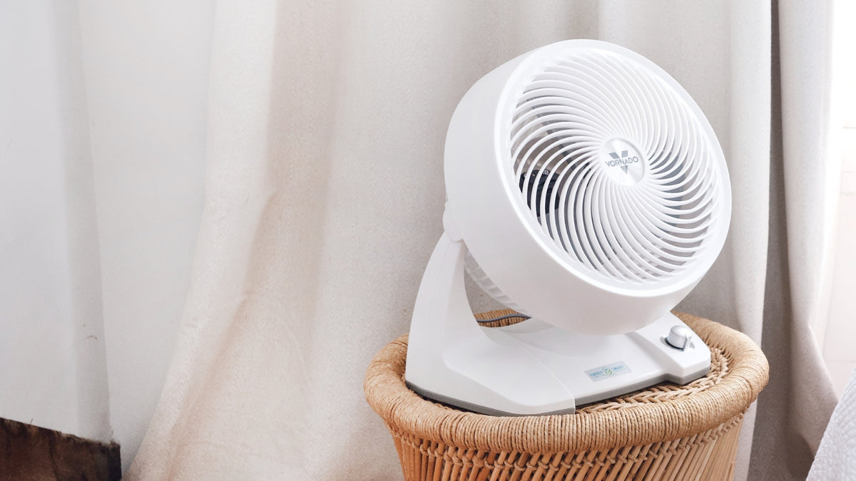 A white Vornado 633DC Energy Smart Air Circulator fan with a ribbed front grille sits on a woven wicker basket in front of soft beige curtains.