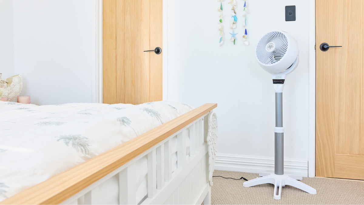 A white Vornado Pedestal Air Circulator standing on a beige carpeted floor next to a bed in a bright bedroom with light wood doors and white wall.