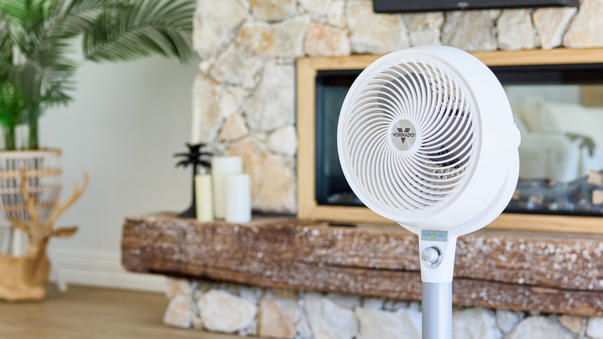 A white Vornado Pedestal Air Circulator positioned in front of a stone feature wall in a living room setting.