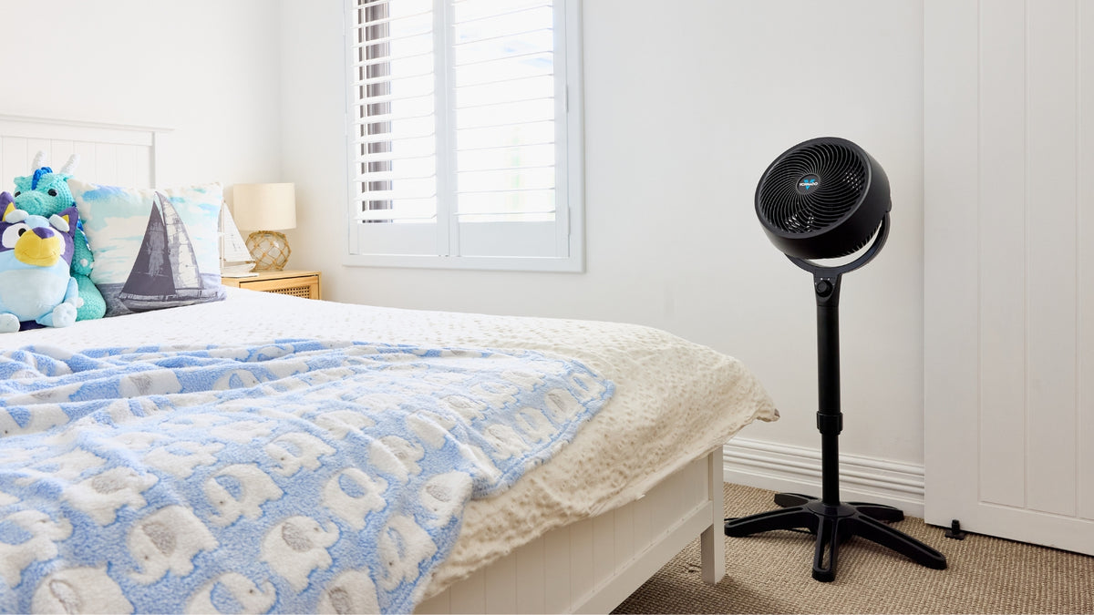 A black Vornado Air Circulator  positioned next to a bed in a bright, modern bedroom with white walls and a window.