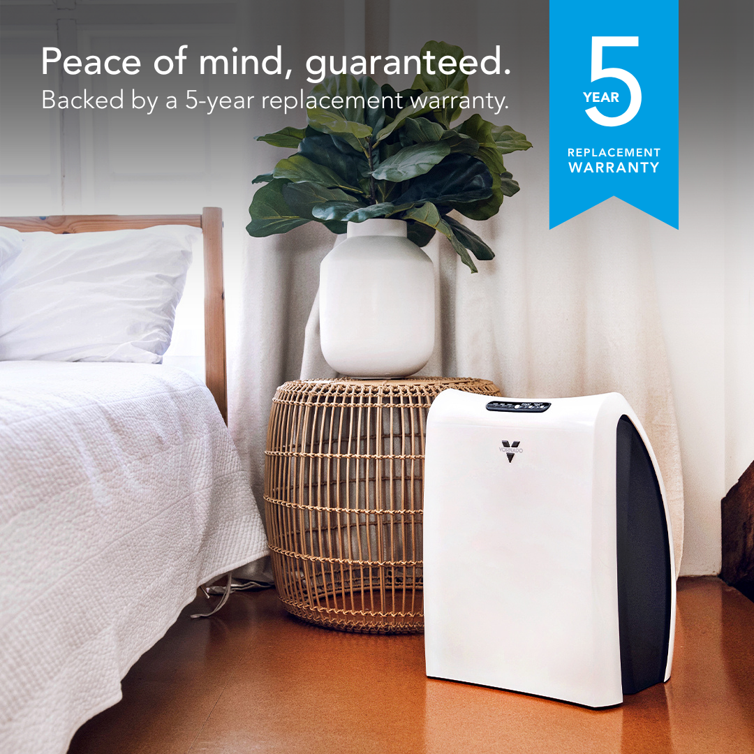 A white and black air purifier placed on a wooden floor in a bedroom setting next to a bed and a wicker side table with a potted plant. Text on the image reads 'Peace of mind, guaranteed.
