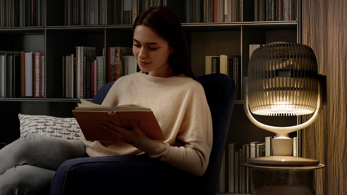 A woman sits in a dark armchair reading a book in a cozy library or study, with a modern Vornado EOS 9 Oscillating Circulator Fan in lamp mode glowing on a side table beside her. Bookshelves fill the background.