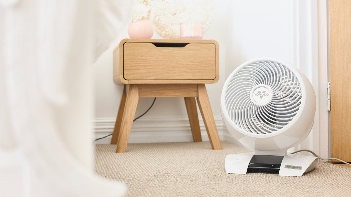 A white and silver Energy Smart™ Air Circulator sitting on a beige carpet next to a light wood nightstand in a bright, modern bedroom setting.
