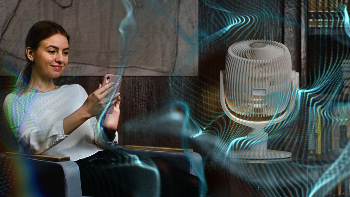 A woman in a white top sits in a chair holding a phone, with a white Vornado EOS 9 Oscillating Circulator Fan is displayed prominently on a side table to the right. The scene has a stylized, digitally edited look with glowing blue wave overlays that suggest airflow and a dark interior background.